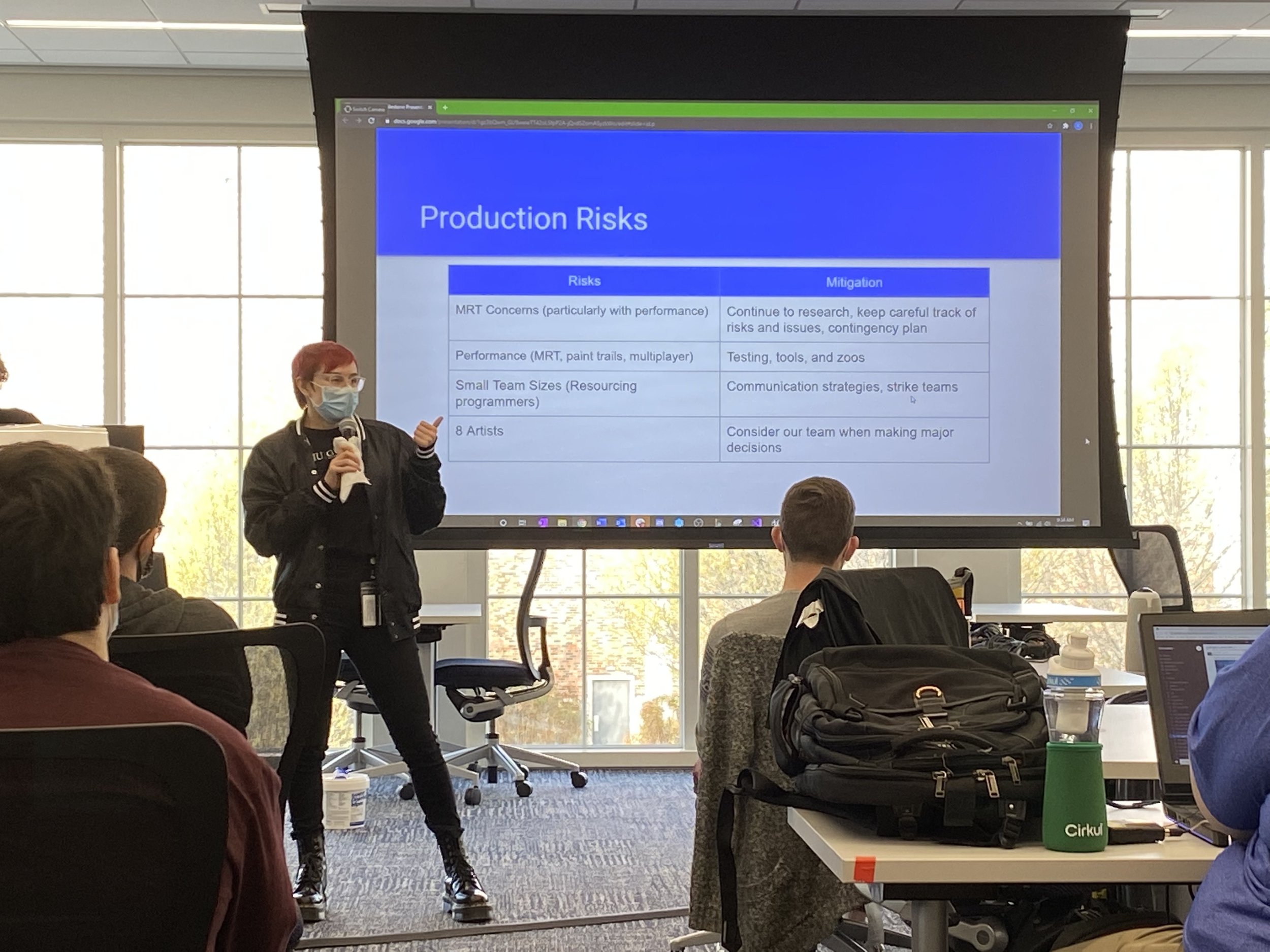 Photograph of Brooke holding a microphone, giving a presentation. On screen behind them is a table going over risks and mitigations.
